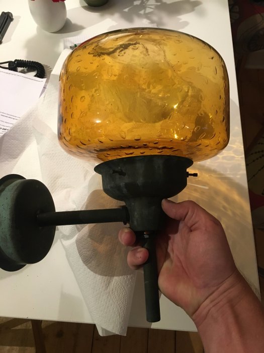 En person håller en gammal vägglampa med gul bubblig glasskärm och svart armatur.