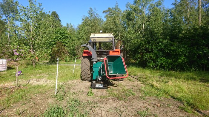 Traktor med monterad flishugg parkerad på en gräsbevuxen yta omgiven av träd.