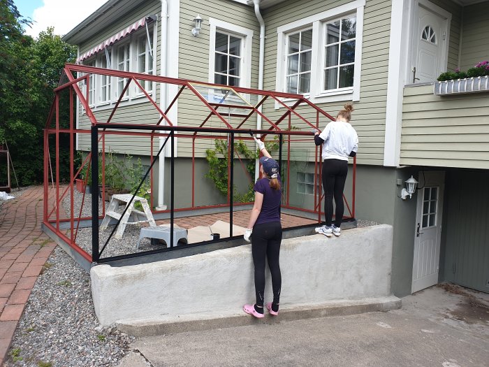 Rödmetallisk växthusram monteras på betonggrund intill hus, med två personer som arbetar.