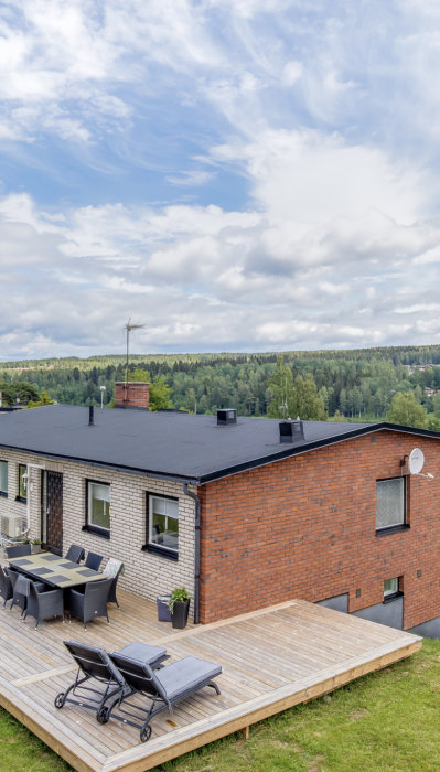 Flat takkonstruktion på ett tegelhus med en terrass och möbler i förgrunden under en himmel med moln.