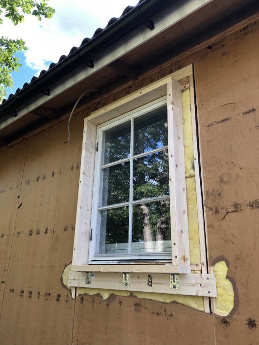 Nyinstallerad fönsterram monterad på reglar med förstärkta vinkeljärn och delvis synlig isolering på en husvägg.