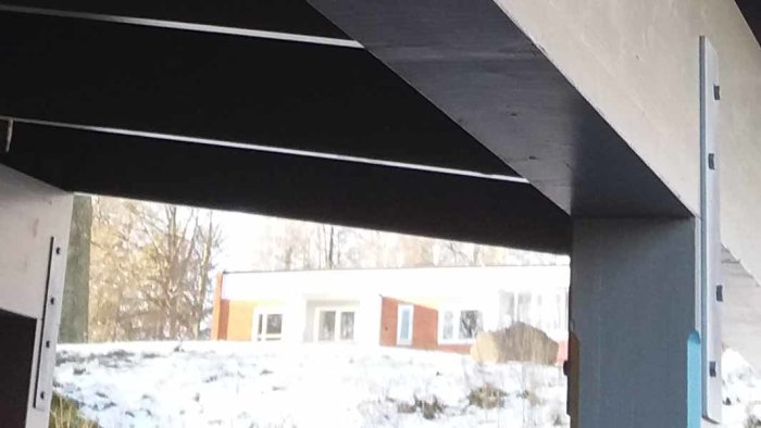 Vy från under ett svart tak mot en vit byggnad med orange detaljer, omgiven av ett vinterlandskap.