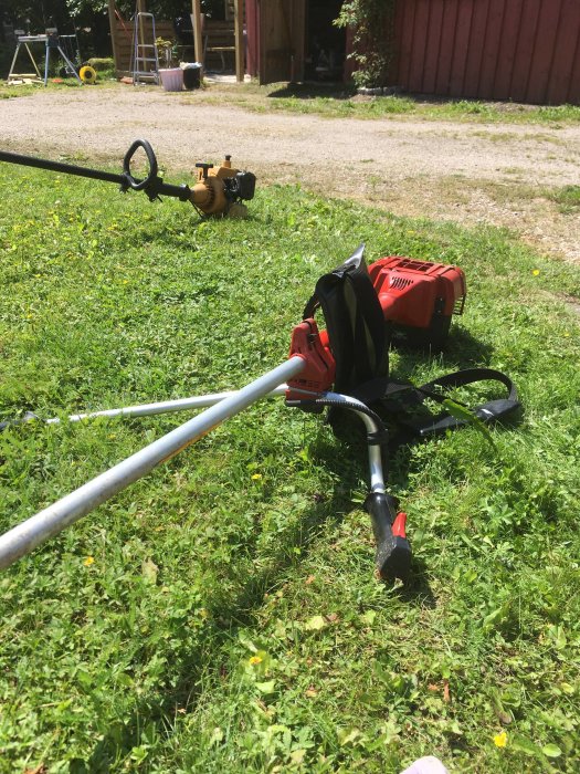 Två bensindrivna trimmers liggande på gräs med ladan i bakgrunden.