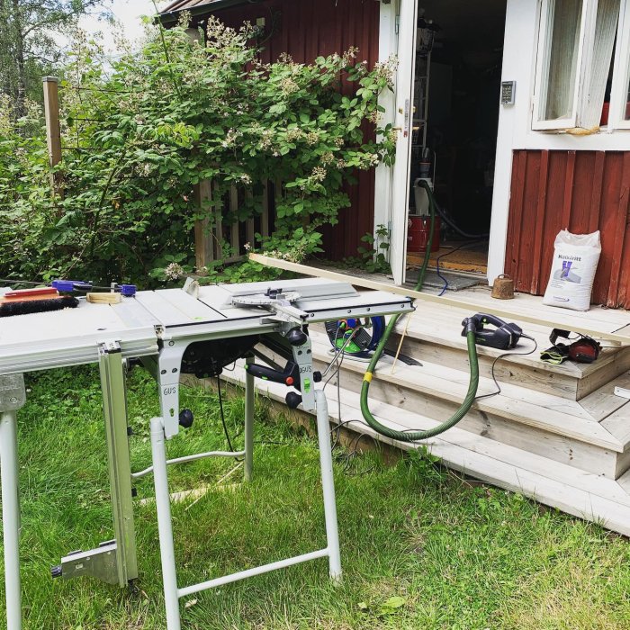 Byggplats med en bordsåg framför en halvfärdig trätrapp eller ramp vid en rödmålad byggnad omgiven av grönska.