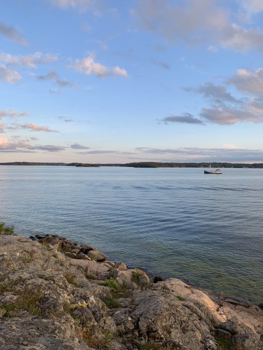 Klippig strandlinje med lugnt vatten och en båt under en molnig himmel som antyder omväxlande väder.