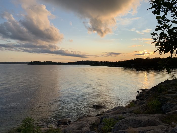 Solnedgång över en sjö med dramatiska moln, solljus som reflekteras i vattnet och en klippig strandlinje.