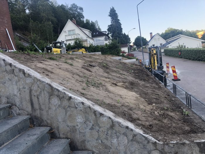 Osäkrad jordig slänt vid hus och gata, redo för trädgårdsarbete med dekorsten och växter.