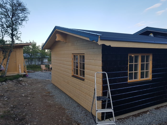 Nybehandlad östfasad på ett trähus med tjära, figursågad nockbalk, stegar vid vägg.