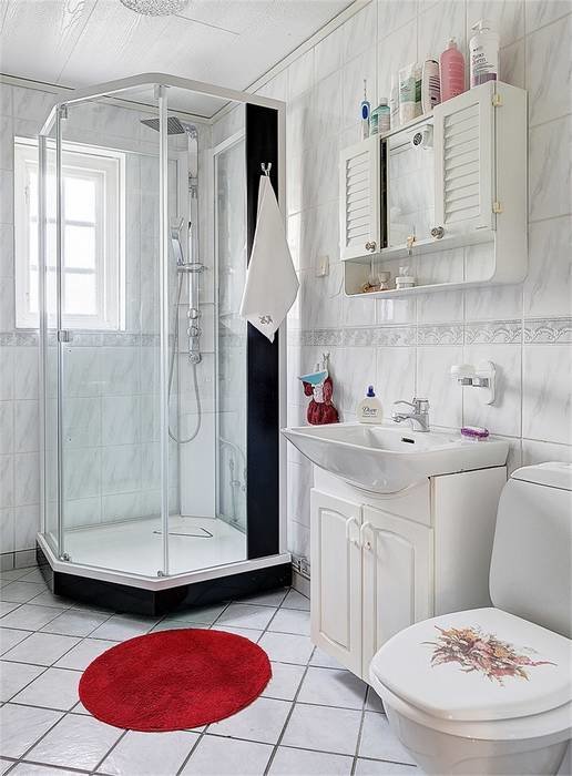 Vit kaklad badrum med duschhörna, toalett, handfat, och röd matta.