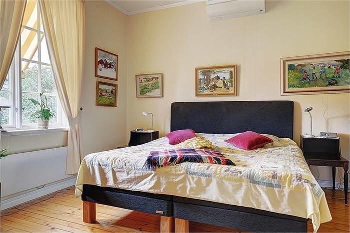 Sovrum med trägolv, stor säng, tavlor på väggen och fönster med gröna växter.