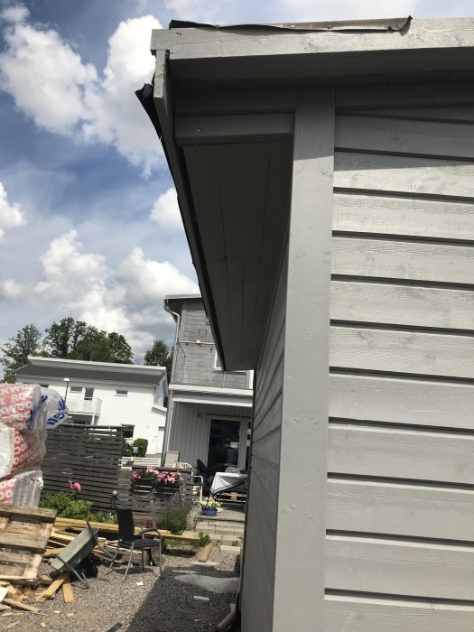 Nymålad grå träkonstruktion med taköverhäng, mot bakgrund av en trädgård och ett större hus.