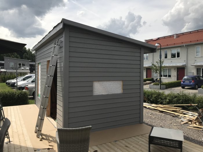 Nymålat grått friggebod under konstruktion på trädäck med stege och byggmaterial runtomkring.