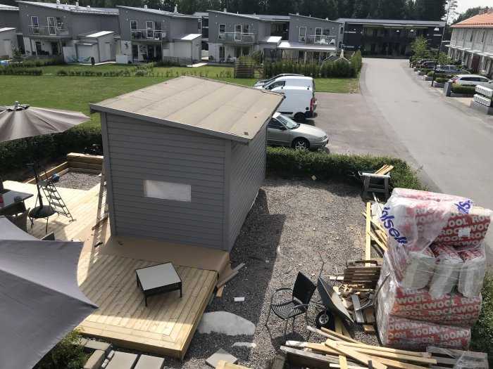 Nymålat litet uthus på träaltan i bakgård, byggmaterial runtomkring, med bostadshus i bakgrunden.