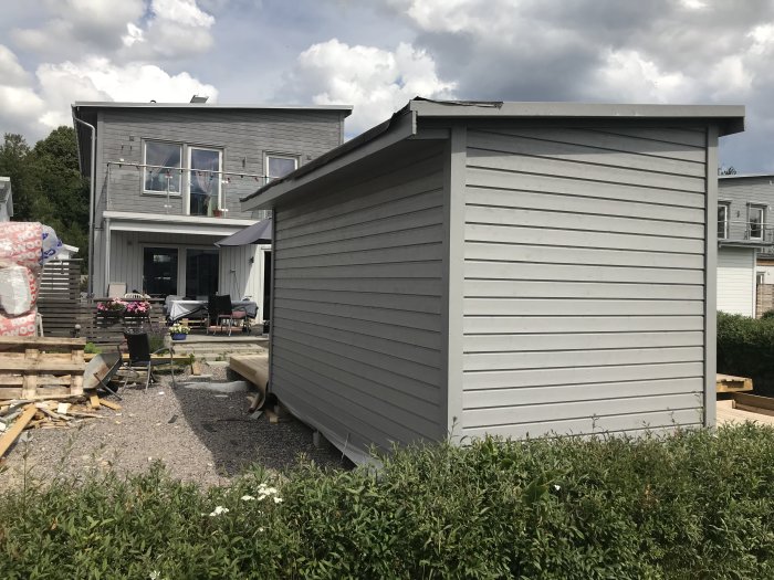 Nymålat trädgårdsskjul i grått som matchar huvudbyggnaden i bakgrunden, väntar på fönster och dörr.