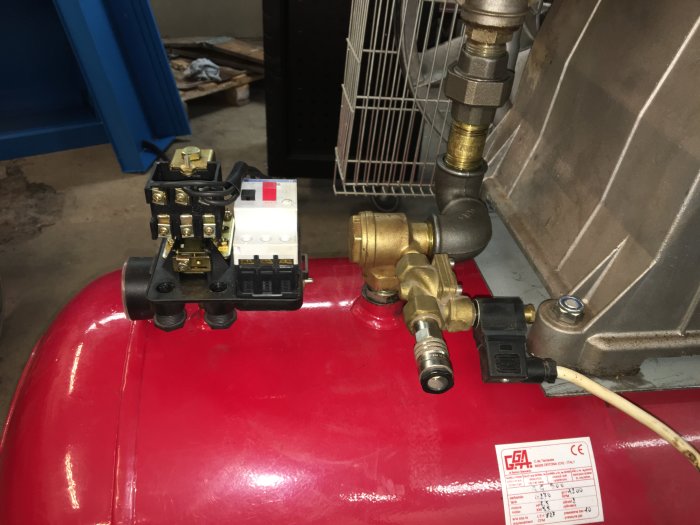Mekanisk utrustning på röd tank med kopplingar, slangar och elektriska komponenter i ett verkstadsutrymme.