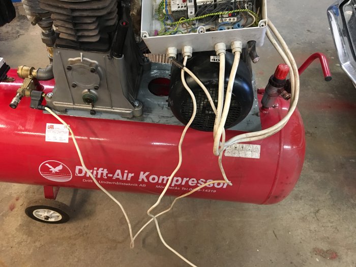 Röd luftkompressor med påmonterad motor och sladdar i ett verkstadsutrymme.