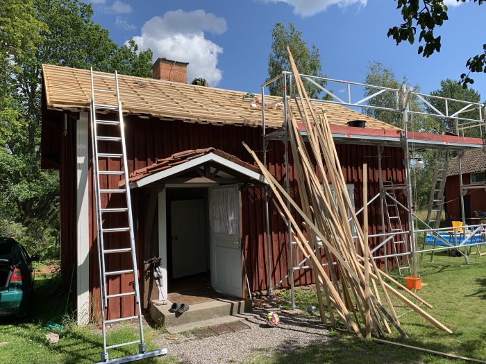 Nykläckt tak på ett rött torp med synligt träarbete, stegar och byggnadsställningar under en solig himmel.