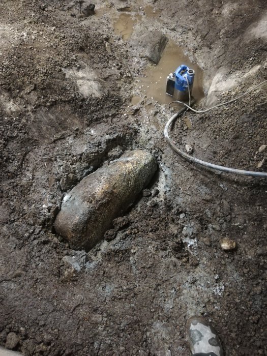 Uppgrävt källargolv med stor sten, grus och lera, inklusive en pump och slangar, under pågående renoveringsarbete.