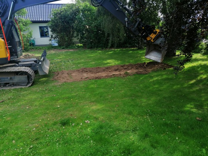 Grävmaskin vid brunnsgrävning på en gräsmatta, uppgrävd jord syns och ett hus i bakgrunden.