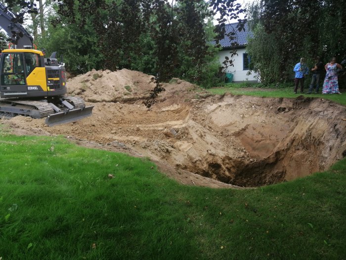 Grävmaskin bredvid en stor brunnsgrävning, ca 6,5 meter djup, i en trädgård med åskådare.