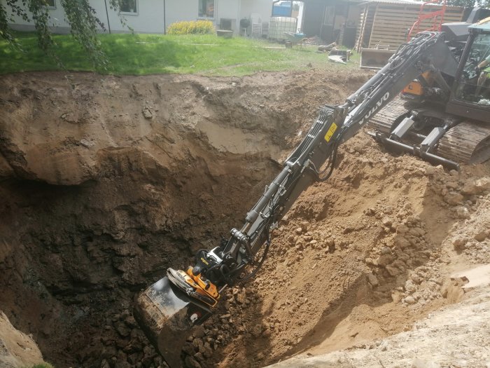 Grävmaskin från Volvo gräver en stor grop i en trädgård för ett byggprojekt.