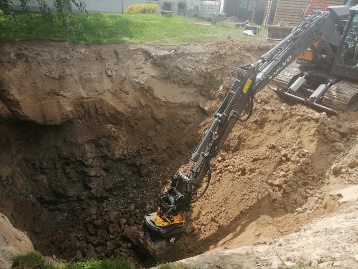 Grävmaskin gräver en stor grop i en trädgård för ett byggprojekt.