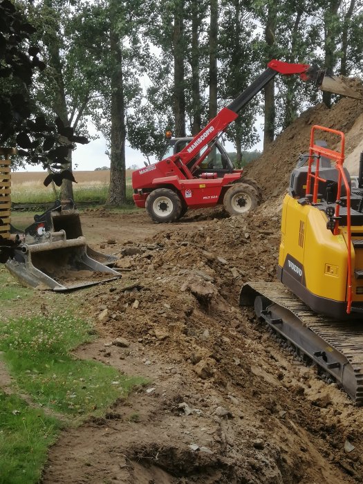 Röd teleskoplastare och gul minigrävare vid arbete på en jordig byggplats med träd i bakgrunden.