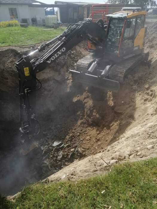 Volvo grävmaskin i arbete med att gräva en stor grop i en trädgård, jord och sten syns.