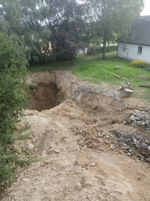 Stor grävd grop i trädgård vid enstaka hus, med grushög och grävmaskinskopa i förgrunden.