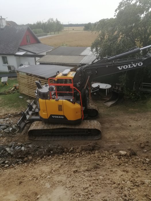 Gul Volvo grävmaskin på band vid ett pågående markarbete nära ett hus med röda takdetaljer och fält i bakgrunden.