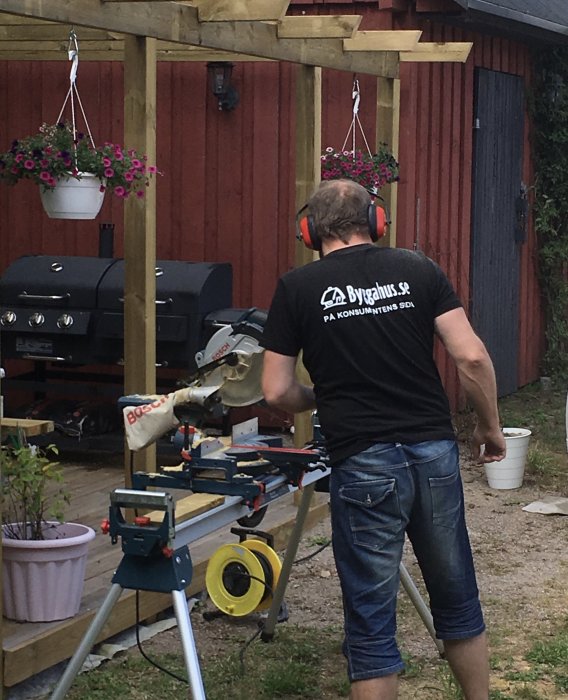 Man i svart t-shirt använder kapmaskin vid utomhusrenovering nära pergola och grill.