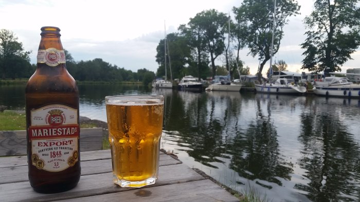 Flaska och glas med öl på träbrygga framför en småbåtshamn.