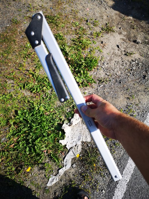 En hand håller en hopfällbar såg med tre blad mot marken med gräs och grus.