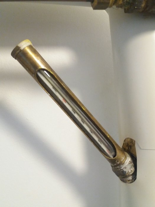 Gammal trasig termometer monterad på ett värmesystems rör med synlig lindad mutter.