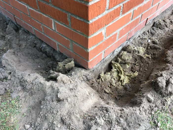 Husgrund med ett nyligen grävt dike som visar isolering 15 cm under jordytan bredvid tegelvägg.