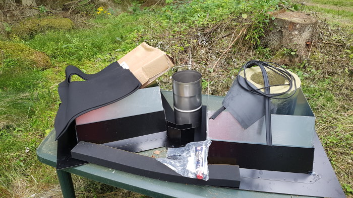 Olika svarta delar och tätningar för kamin, bl.a. ett nockbeslag, på ett grönt bord utomhus.