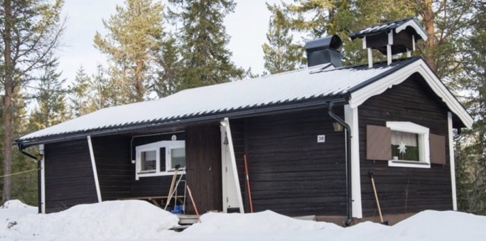 Svartmålad stuga med snöig altan under renovering, stege och snöhög framför.