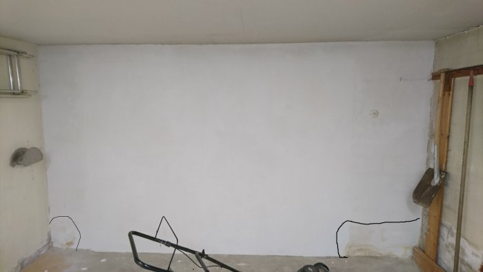 Vitmålad källarvägg med igenmurad garagedörr och svarta markeringar runt fuktfläckar nära golvet.