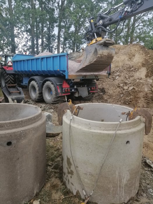 Grävmaskin tömmer grus i en traktorkärra nära betongbrunnar under sättning och återfyllning.