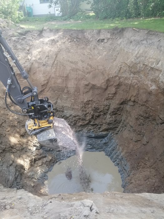 Grävmaskinsarm häller grus i ett stort hål med vatten under byggprojektet.