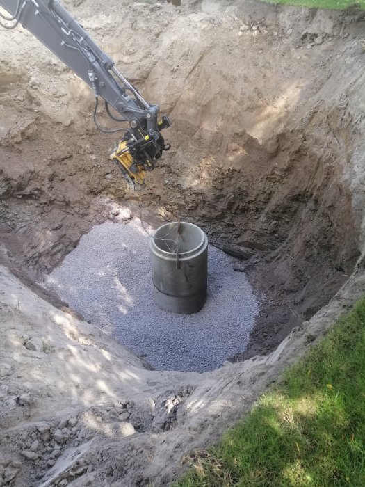 Grävmaskinsarm som sätter ner en brunn i en grävd grop med grusunderlag.