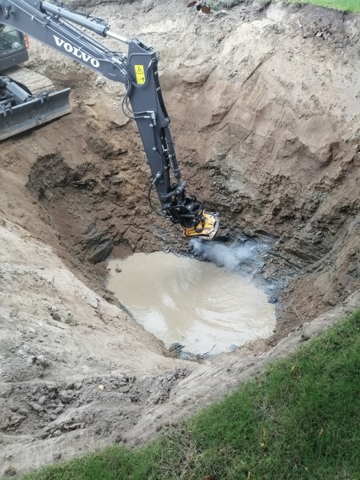 Grävmaskin från Volvo som arbetar i en grävd brunn med vatten och återfyllning runt sidorna.