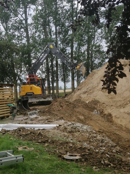 Grävmaskin av märket Volvo i arbete på en byggarbetsplats med en hög grus och rörledningar på marken.