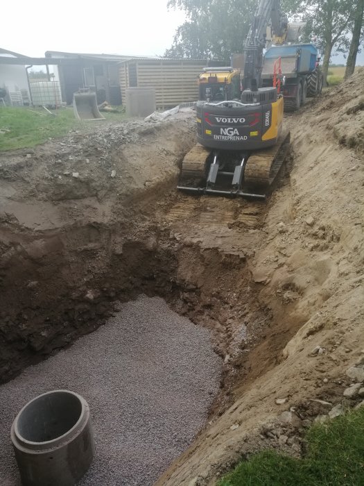 Grävmaskin från Volvo vid en byggarbetsplats gräver ut en grop fylld med grus bredvid en betongbrunn.