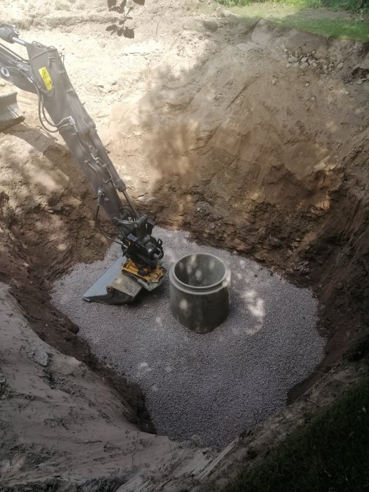 Grävmaskinsarm i utgrävd grop med grus och betongbrunn för grundarbeten.