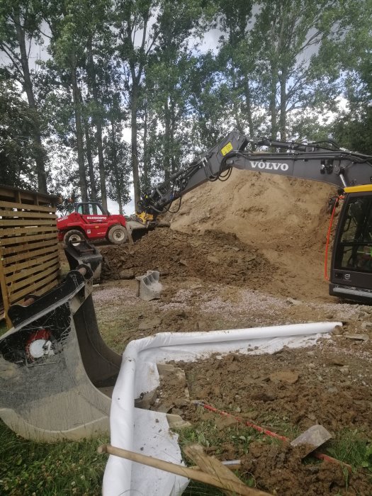 Grävmaskin från Volvo och teleskoplastare i arbete vid en byggplats med upphöjd jordhög och byggmaterial i förgrunden.