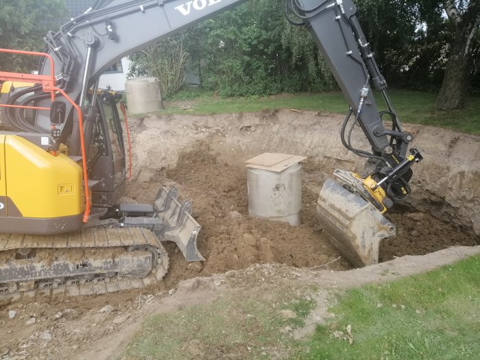 Minigrävare gräver en grop i trädgård nära betongringar, sannolikt för avlopp eller brunnar.