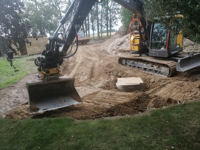 Kompakt grävmaskin gräver i lerig terräng, omgivet av grön vegetation och en staplad trästaket i bakgrunden.