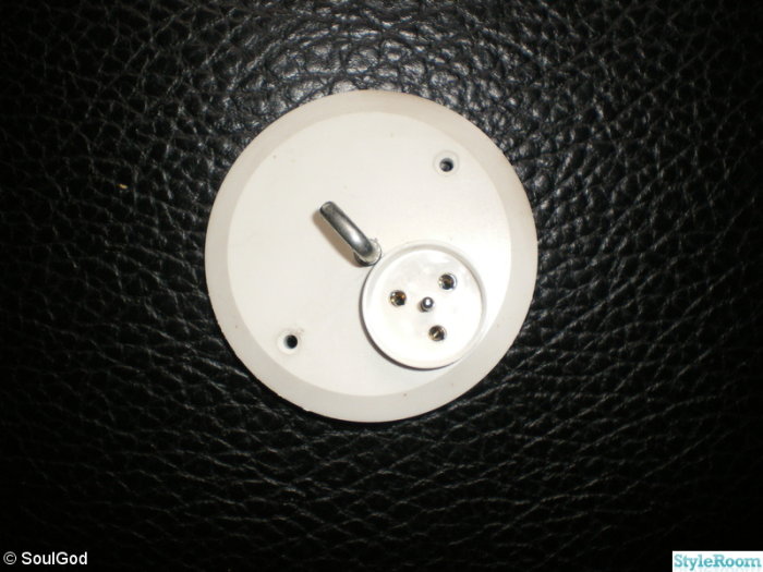 Vit kronlamputtag med ett centrumjordstift och tre hål, monterad på en mörk texturerad yta.