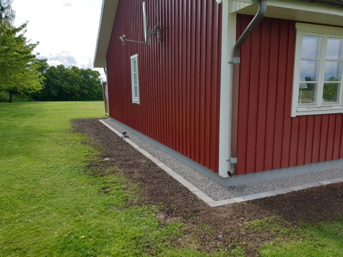 Renoverad husgrund med nytt lager makadam och marksten vid rödmålat hus.